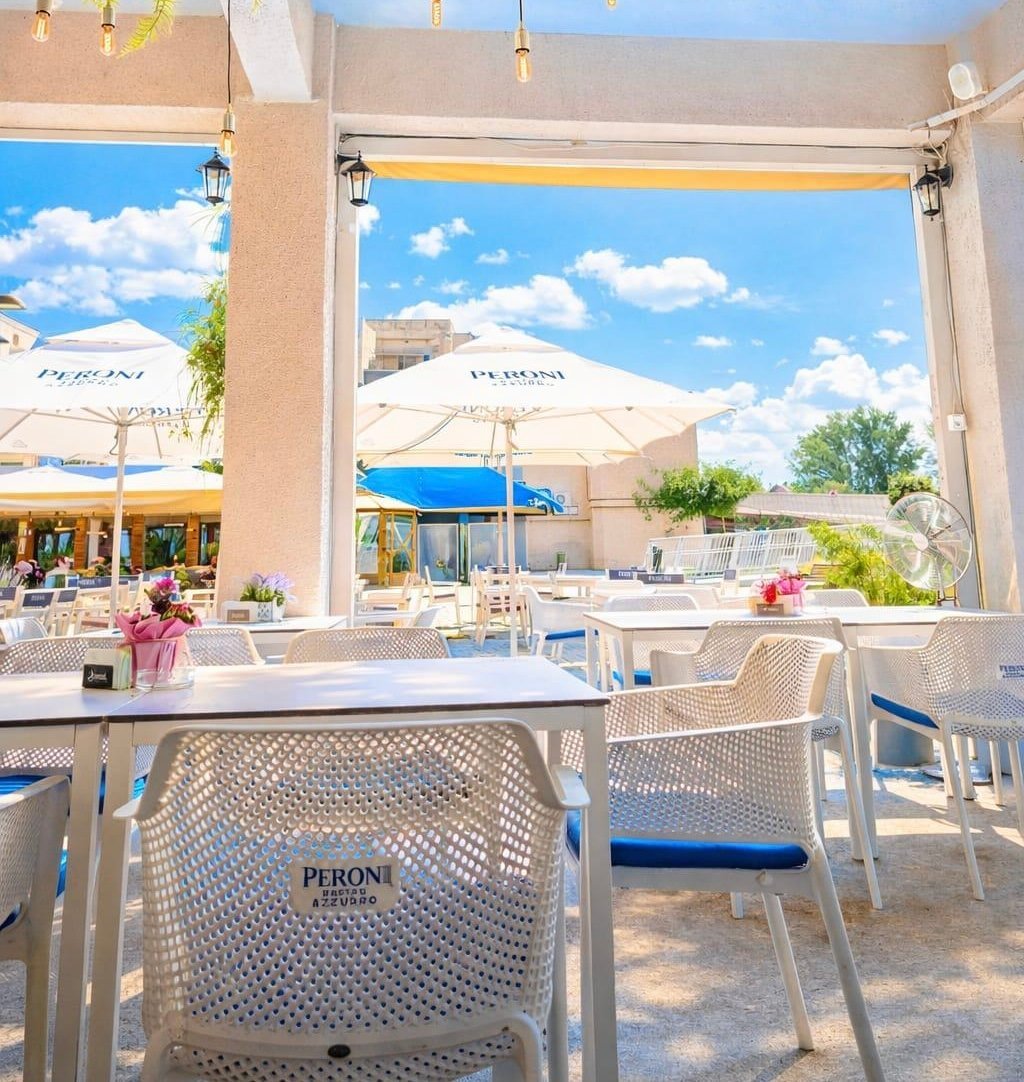Outdoor restaurant patio with white Peroni chairs, tables, and umbrellas overlooking a blue sky and landscape
