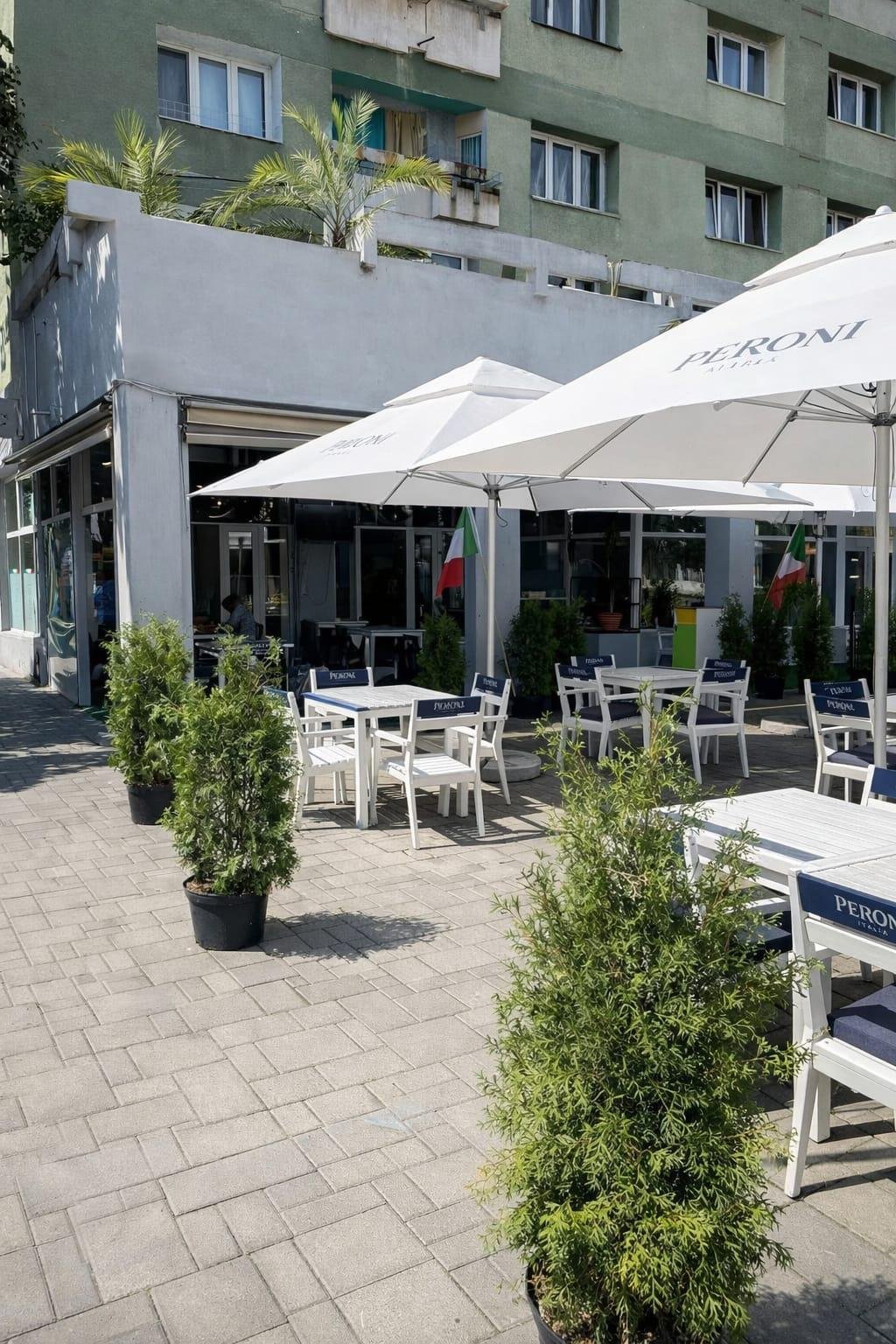 Outdoor restaurant patio with white umbrellas, dining tables and chairs, green plants, modern storefront and residential building